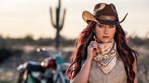 Woman in cowboy hat and leather vest standing by motorcycle, crank it up by Ashley Wineland.