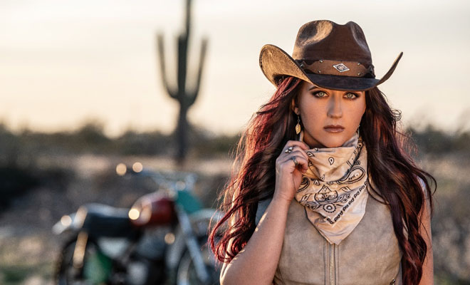 Woman in cowboy hat and leather vest standing by motorcycle, crank it up by Ashley Wineland.