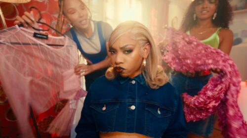 Three women in a brightly lit room. The woman in the center, wearing a denim jacket, has a pouty expression. The other two, vibing to "Controllla by Fave," are holding up pink and purple garments while looking at her.