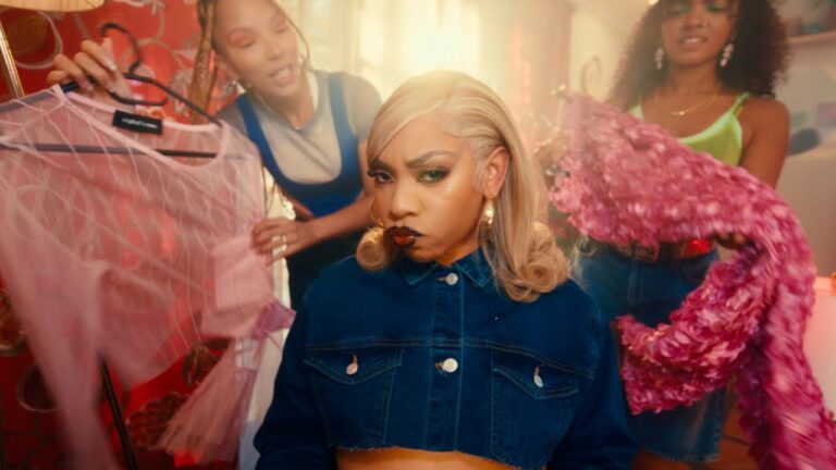 Three women in a brightly lit room. The woman in the center, wearing a denim jacket, has a pouty expression. The other two, vibing to "Controllla by Fave," are holding up pink and purple garments while looking at her.