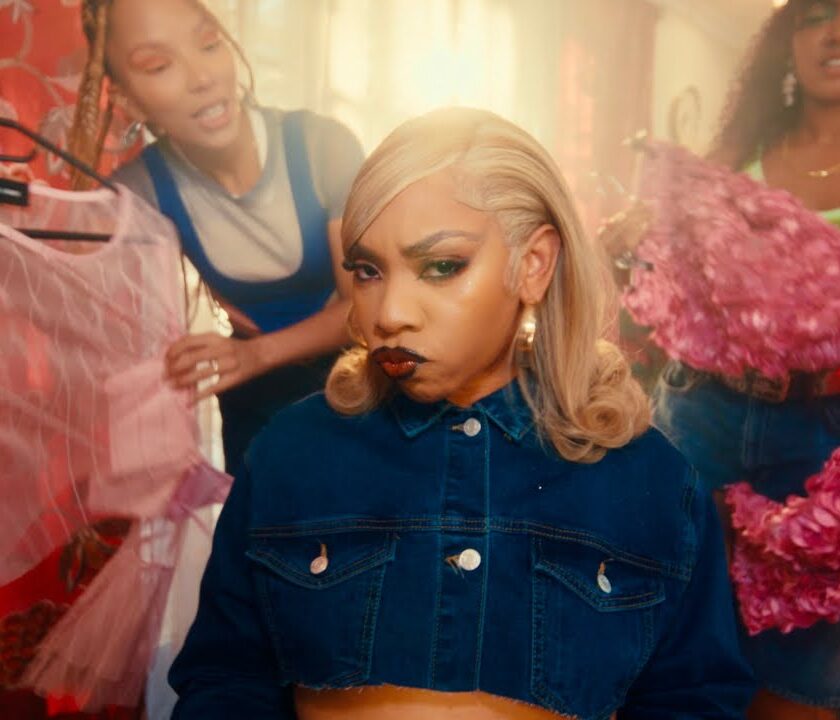 FAVE – Controlla (Official Music Video) Three women in a brightly lit room. The woman in the center, wearing a denim jacket, has a pouty expression. The other two, vibing to "Controllla by Fave," are holding up pink and purple garments while looking at her.