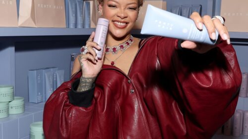 A person in a red outfit smiles while holding two hair product bottles in a store with shelves stocked with similar products.
