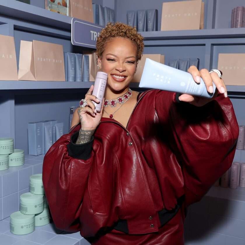Rihanna Stuns with Natural Curls at Fenty Hair Launch Party A person in a red outfit smiles while holding two hair product bottles in a store with shelves stocked with similar products.