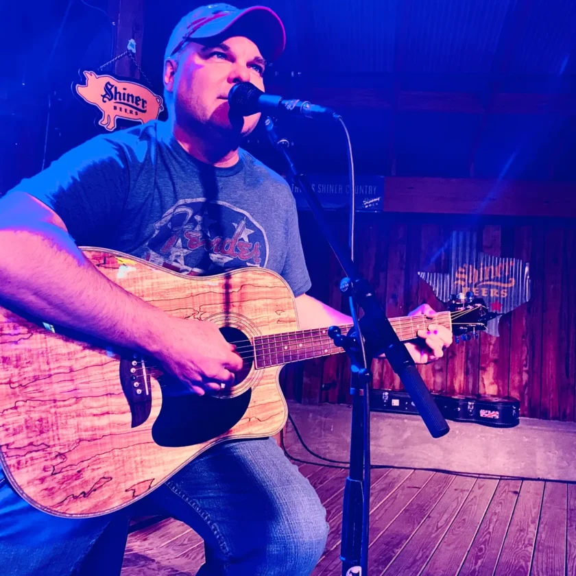A man wearing a cap and t-shirt plays an acoustic guitar and sings into a microphone on a small wooden stage under blue lighting.